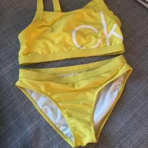 Gul bikini från Calvin Klein - Snygg gul bikini från Calvin Klein med sportig topp och klassiska bikiniunderdelar. Toppdelen har vita CK-bokstäver framtill och breda axelband. Underdelen har Calvin Klein-logga baktill. Materialet är mjukt och stretchigt syntet 💗