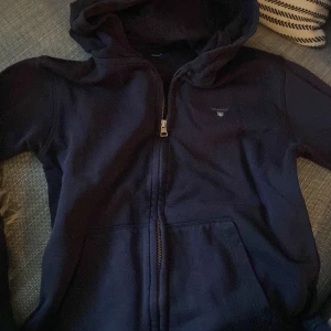 Marinblå hoodie från GANT - Mörkblå hoodie från GANT med dragkedja framtill och broderad logga på bröstet. Tröjan har huva och två fickor framtill. Perfekt för en avslappnad stil och funkar året runt. 