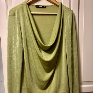 Ljusgrön omlottopp från Bik Bok - Säljer en ljusgrön omlottopp med urringning från Bik Bok i storlek XL. Toppen har en snygg draperad front med djup omlottringning och långa ärmar. Materialet är mjukt och skimrande syntet som ger en  glansig känsla.