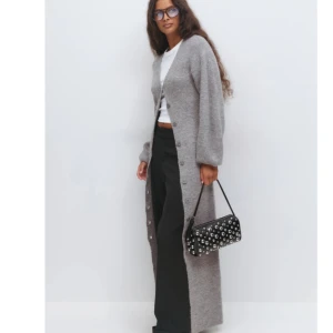 Lång grå stickad väst med knappar - Lång, mysig grå stickad väst med v-ringning och knappar hela vägen framtill. Västen har en avslappnad passform och ballongärmar för en trendig look. Perfekt att slänga över en t-shirt för en chill vibe.