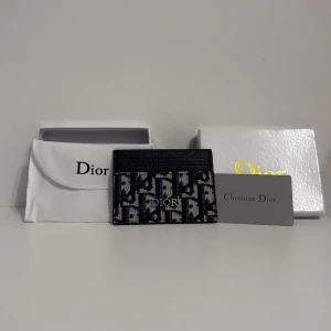 Svart och vit Dior plånbok - Snygg plånbok från Dior med klassiskt monogrammönster i svart och vitt. Plånboken har en stilren och kompakt design med flera fack för kort. Materialet är en mix av skinn och textil, och loggan i silver ger en lyxig touch. Perfekt för dig som gillar exklusiva accessoarer.