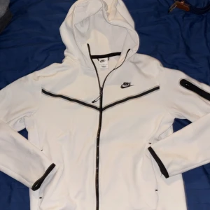 Vit Nike tech med huva, M - Nike tech i vitt med svarta detaljer och dragkedja framtill. Jackan har huva, lång ärm och en snygg svart linje över bröstet. Fickor med dragkedja och logga på vänster sida. Perfekt för chill och streetwear.
