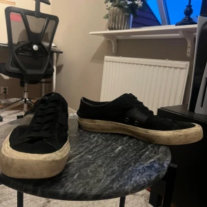 Svarta sneakers från Tom Ford - Svarta sneakers från Tom Ford med klassisk låg profil och rund tå. Skorna har snörning och en beige, något smutsad platt sula. Materialet är mocka med detaljer i skinn och logga på hälen. Perfekta för dig som gillar stilrena och exklusiva sneakers.