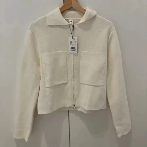 Vit kofta med dragkedja från Åhlens - Snygg vit kofta från Åhlens i storlek XS. Modellen är croppad med två stora fickor framtill, klassisk krage och hel dragkedja. Tillverkad i ett stickat material som känns mjukt och bekvämt. Perfekt för lager-på-lager under kyliga dagar.