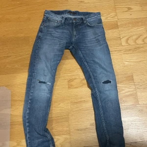 Nudie jeans - Snygga blå Nudie jeans med skinny passform och slitningar på knäna. Klassisk femficksmodell med läderpatch bak och dragkedja i gylfen. Jeansen har en tvättad look och är gjorda i stretchigt denim för skön komfort.
