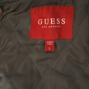 Grå och svart bomberjacka från Guess - Säljer en grå och svart bomberjacka från Guess med coola broderade detaljer och patchar. Jackan har svarta skinnärmar, ribbade muddar och knappar framtill. Insidan är quiltad och jackan har en avtagbar svart hoodie. Perfekt för dig som vill ha en streetig look.