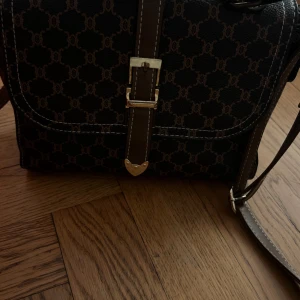 Brun axelväska  - Snygg brun axelväska  med klassiskt mönster och guldfärgade detaljer. Väskan har justerbar axelrem, magnetlås och dragkedja på insidan. Perfekt storlek för det viktigaste och stilren design med kontrastsömmar.