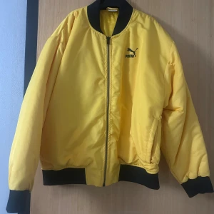 Gul bomberjacka från Puma - Säljer en fet gul bomberjacka från Puma med svart logga på bröstet och texten 'Automne-Hiver' i svart på ryggen. Jackan har svarta muddar och krage, dragkedja framtill och två sidofickor. Perfekt statement-plagg för dig som gillar streetstyle.