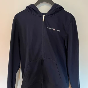 Marinblå hoodie från Gant med dragkedja - Snygg marinblå hoodie från Gant med dragkedja framtill och broderad logga på bröstet. Tröjan har huva, känguruficka och är tillverkad i mjuk bomull. Perfekt för en chill och sportig stil.
