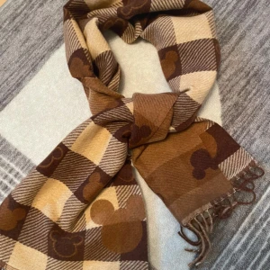 Brun rutig halsduk med Musse Pigg-mönster - Snygg halsduk i rutigt mönster med olika nyanser av brunt och beige. Halsduken har Musse Pigg-huvuden invävda i rutorna och fransar i ändarna. Perfekt accessoar för att ge din outfit en lekfull touch.