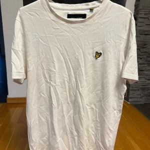 Vit t-shirt från Lyle & Scott - Vit t-shirt från Lyle & Scott med klassisk passform och korta ärmar. På bröstet sitter den ikoniska gula fågeln som broderad detalj. Materialet är mjuk bomull och t-shirten har en rund halsringning. Perfekt för en clean och enkel stil.