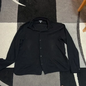 Svart skjorta från H&M XL - Svart skjorta från H&M i regular fit med klassisk krage och knappar framtill. Materialet har en lätt textur som ger en cool känsla. Långärmad modell med knapp vid ärmslut. Perfekt för dig som gillar stilrena och enkla plagg.