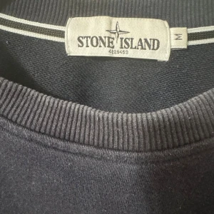 Mörkblå sweatshirt från Stone Island - Säljer en mörkblå sweatshirt från Stone Island i storlek M. Tröjan har klassisk rund hals, ribbade muddar och den ikoniska logotypen på vänster ärm. Perfekt för dig som gillar streetwear och vill ha en clean look. Ganska sliten dock därav billigt pris