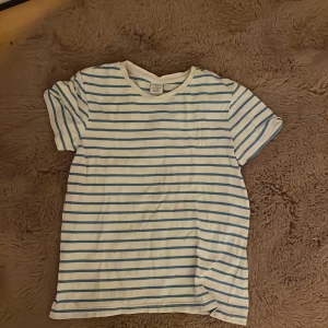 Randig t-shirt från Lindex, blå/vit - Snygg t-shirt från Lindex i storlek 134/140. T-shirten är vit med tunna blå ränder, har rund hals och korta ärmar. Tillverkad i mjuk bomull, perfekt för en avslappnad stil. Klassisk och enkel design som funkar till det mesta.