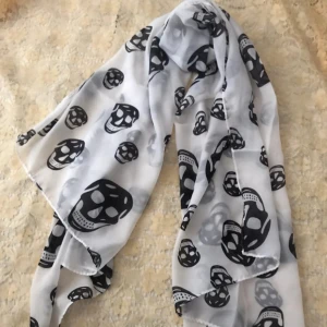 Vit halsduk med svarta döskallar - Snygg och tunn halsduk i vitt med svarta döskallemönster. Perfekt accessoar för dig som gillar lite edgy stil. Halsduken är rektangulär och gjord i ett lätt, transparent material som känns skönt mot huden. Alexander McQueen inspirerad. Mått: 167x70cm