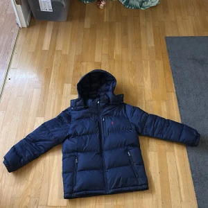 Mörkblå pufferjacka från Ralph Lauren - Snygg mörkblå pufferjacka från Ralph Lauren med huva och dragkedja framtill. Jackan har två sidofickor med dragkedja och den klassiska röda Polo-loggan broderad på bröstet. Perfekt för kalla dagar och har en clean, sportig vibe.