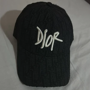 Svart Dior keps med broderad logga - Snygg svart keps från Dior med broderad vit logga framtill. Kepsen har ett diskret mönster med Dior-text över hela ytan och justerbar rem baktill. Materialet är bomull med mesh på insidan för extra komfort. Perfekt för dig som vill ha en exklusiv streetstil.