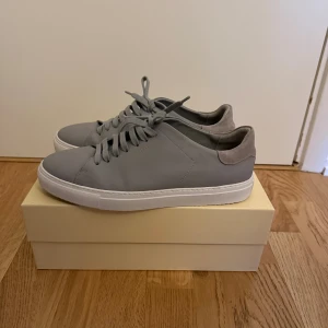 Grå sneakers från Axel Arigato - Snygga grå sneakers från Axel Arigato med stilren design. Skorna har ovandel i slätt skinn och detaljer i mocka vid hälen. Platt vit sula och rund tå. Klassisk snörning framtill och diskret logga på sidan. Perfekta för dig som gillar minimalistisk stil.