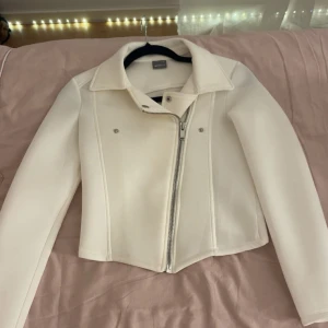 Vit croppad bomberjacka med dragkedja - Snygg vit bomberjacka från gina Tricot med croppad passform och asymmetrisk dragkedja framtill. Jackan har klassisk krage, långa ärmar och stilrena detaljer i silver. Perfekt för dig som gillar minimalistisk och trendig stil.