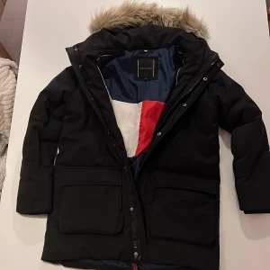 Svart parkajacka Tommy Hilfiger - Parkajacka från Tommy Hilfiger i svart med stor huva och fuskpäls. Insidan har ett snyggt blockmönster i blått, vitt och rött. Jackan har två stora fickor framtill, dragkedja och tryckknappar. Perfekt för kalla dagar och riktigt stilren.