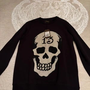 Svart sweatshirt med döskalle Ed Hardy - Svart crewneck sweatshirt från Ed Hardy med stor glittrig döskalle och siffran 13 framtill. Tröjan har långa ärmar och är dekorerad med diamantliknande detaljer som ger en riktigt edgy vibe. Perfekt för dig som gillar statement-plagg och vill sticka ut.