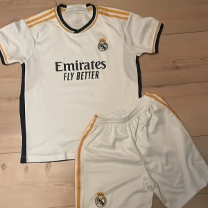 Real Madrid matchställ Adidas vit/gul - Säljer ett Real Madrid matchställ från Adidas med kortärmad tröja och shorts. Setet är vitt med gula och svarta detaljer, klubbmärke broderat på både tröja och shorts samt 'Emirates Fly Better' tryck på bröstet. Materialet är lätt och andas, perfekt för fotbollsträning.
