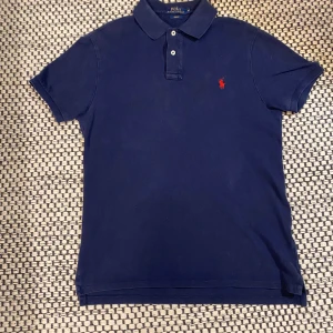 Marinblå pikétröja från Ralph Lauren - Snygg marinblå pikétröja från Polo Ralph Lauren i slim fit-modell. Klassisk design med krage, knappar och röd broderad logga på bröstet.                                         Storlek-M