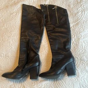 Svarta höga stövlar med klack - Snygga svarta stövlar i skinn med kraftig blockklack. Stövlarna går till knäet och har dragkedjor både på insidan och upptill för en cool detalj. Perfekta för att lyfta vilken outfit som helst.