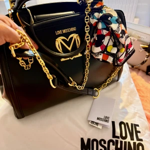 Svart handväska från Love Moschino - Stilren svart handväska från Love Moschino med guldfärgade kedjedetaljer och logotyp framtill. Väskan har en färgglad scarf knuten vid handtaget och är tillverkad i slätt skinn. Rymlig insida och elegant form, perfekt för dig som gillar statement-accessoarer.