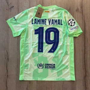 Barcelona Lamine Yamal fotbollströja - Säljer en Barcelona fotbollströja med Lamine Yamal och nummer 19 på ryggen. Tröjan är ljusgrön med mönstrade detaljer, har korta ärmar och är tillverkad i lätt polyester. Officiella klubbmärken, Champions League-patch och sponsorloggor från Spotify och UNHCR finns på tröjan.