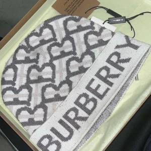 Burberry mössa med hjärtmönster - Exklusiv stickad mössa från Burberry i ljusgrått och vitt med grått hjärtmönster och stor logga framtill. Snygg och trendig accessoar som lyfter din vinterstil. Tillverkad i mjukt material för maximal komfort.