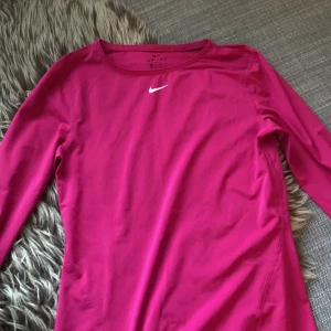 Rosa långärmad Nike Dri-Fit träningströja - Nike Dri-Fit långärmad träningströja i stark rosa färg. Tröjan har rund halsringning, diskret Nike-logga på bröstet och är tillverkad i ett lätt, ventilerande syntetmaterial som andas. Perfekt för löpning eller gympass. Kontakta innan köp!!