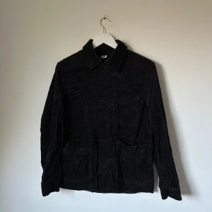 Svart manchester overshirt från Arket - Svart overshirt/jacka från Arket i mjuk manchester med klassisk krage, stora fickor framtill och knäppning med knappar. Perfekt för lager-på-lager och har en clean, stilren look som funkar till många outfits. Materialet är en bomullsblandning. 4F14