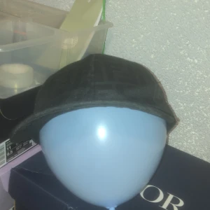 Svart keps från Fendi med monogram - Säljer en svart keps från Fendi med diskret monogrammönster över hela kepsen. Klassisk böjd skärm och justerbar rem baktill med metallspänne. Snygg och stilren accessoar som lyfter din outfit.