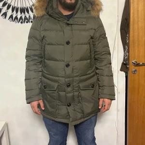 Olivgrön Moncler dunjacka med päls - Säljer en olivgrön Moncler dunjacka med fluffig päls på huvan, stora fickor med knappar och både dragkedja och knappstängning. Jackan har Moncler-logga på ärmen och coolt serietidningsfoder på insidan. Perfekt för kalla dagar och riktigt snygg passform.
