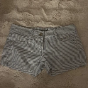 Randiga shorts  - Snygga korta shorts från Lindex med smala blå och vita ränder. Klassisk femficksmodell med knapp och dragkedja framtill. Perfekta för varma dagar och har en fräsch, ungdomlig stil.