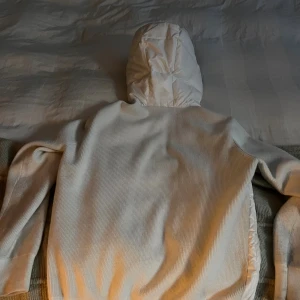 Vit hoodie från Moncler med huva - Säljer en snygg vit cardigan från Moncler med ribbad struktur och puffad huva i glansigt material. Tröjan har långärmade ärmar och Moncler-logga på axeln. Perfekt för dig som gillar exklusiva märken och stilren design.