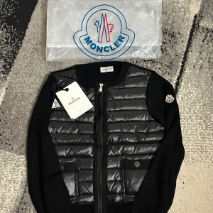 Moncler Cardigan  - Snygg svart pufferjacka från Moncler med quiltad framsida och två fickor med knappstängning. Jackan har rund hals, dragkedja och Moncler-logga på ärmen. Materialet är en mix av polyester och dun, vilket gör den både lätt och varm. Perfekt för dig som gillar stilren design.