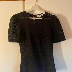 Svart spetsblus från Vila - Supersnygg svart blus från Vila med korta ärmar och vackert spetsmönster över hela toppen. Blusen har rund halsringning och lätt puffade axlar för en trendig look. Perfekt att styla med jeans eller kjol för en cool outfit.