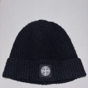 Svart stickad mössa Stone Island - Svart ribbstickad mössa från Stone Island med klassisk logopatch framtill. Snygg och enkel design som passar perfekt för kalla dagar. Mjuk och varm känsla tack vare det stickade materialet.