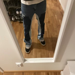 Unika blå cargopants med färgstänk - Säljer ett par riktigt coola flared jeans i olika blå nyanser med vita färgstänk och kontrasterande paneler. Byxorna har flera fickor, raka ben och en loose passform. Perfekt för dig som vill sticka ut med din stil. Materialet är jeans och detaljerna gör dem extra speciella.