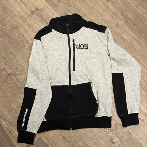 Grå och svart hoodie XL Professions - Snygg grå och svart hoodie från Professions XL med hel dragkedja, huva och praktiska fickor. Har coolt XL-tryck på bröstet och textdetalj på ärmen. Perfekt för chill eller träning, med kontrastpaneler och sportig vibe.