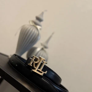 Svart skärp från Ralph Lauren - Snyggt svart bälte från Ralph Lauren med en stilren RL-logga i guld som spänne. Bältet är tillverkat i slätt skinn och har klassisk form med flera hål för justering. Perfekt för att lyfta din outfit med en exklusiv touch.