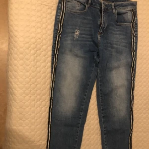Blå jeans med svarta sidoränder - Snygga blå jeans med slitningar och svarta sidoränder med vita detaljer längs benen. Klassisk femficksmodell med dragkedja och knapp. Jeansen har en smal passform och är tillverkade i stretchigt denimtyg för extra komfort.