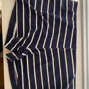 Randiga blå shorts från Lefties - Säljer ett par snygga shorts från Lefties i storlek S men passar XS. De är marinblå med vita vertikala ränder och har en bekväm midwaist-passform. Materialet är mjuk bomull, perfekt för varma dagar. Stilen är casual och passar dig som gillar enkel och fräsch design.