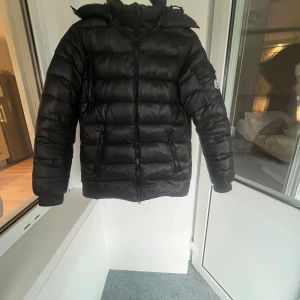 Svart Moncler pufferjacka med huva - Svart pufferjacka från Moncler med huva och två sidofickor med dragkedja. Jackan har Moncler-logga på ärmen och quiltad design. Perfekt för kalla dagar med sin dunfyllda insida och stilrena look. Dragkedja framtill och elastiska muddar vid ärmslut.