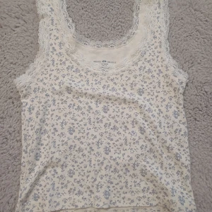 Blommig spetskantad linnetopp Brandy Melville - Supersöt vit linnetopp från Brandy Melville med småblommigt blått mönster och spetskant runt halsringning och ärmhål. Tillverkad i 100% bomull för en mjuk och skön känsla. Perfekt för en romantisk och chill stil.