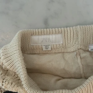Stickade beige byxor från Zara - Säljer ett par beige stickade byxor från Zara med snyggt hålmönster och svarta detaljer vid bensluten.( De är lite för långa för mig som är 161)