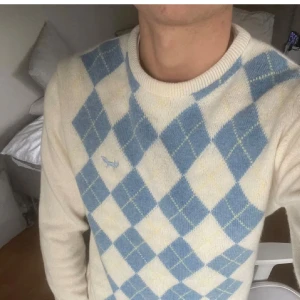 Beige och blå argyle stickad tröja - Snygg stickad tröja med klassiskt argylemönster i beige och ljusblått. Tröjan har rund halsringning, långa ärmar och en liten broderad logga på bröstet. Perfekt för dig som gillar preppy stil och vill sticka ut lite extra.