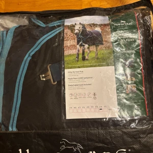 Svart och blå Amigo Bravo hästtäcke - Amigo Bravo 12 Plus Pony hästtäcke i svart med blå detaljer. Täcket är tillverkat av slitstark 1200D polyester, har avtagbar huva och vattentätt material. Passar ponny, medium 250g fyllning. Snygg design med praktiska spännen och andasfunktion.